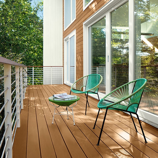 Dos modernas sillas de ratán verde y una mesa en una terraza con piso de madera marrón claro y barandillas blancas, que se extiende desde el exterior de la casa revestida de madera en color marrón claro, marcos pintados de blanco y ventanas del piso al techo.