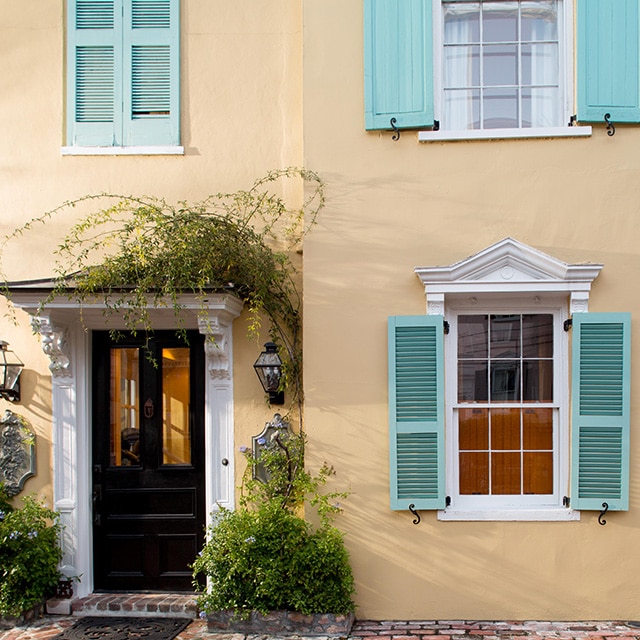 Un bonito exterior de estuco pintado de beige con un suave tono durazno, las contraventanas de color verde azulado, los marcos blancos y la vegetación alrededor de la puerta de entrada negra.