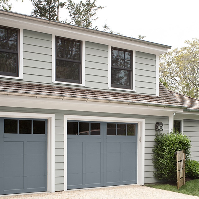 Ces portes de garage grises offrent un contraste joliment équilibré avec le parement blanc cassé aux nuances gris-bleu de cette maison et à ses moulures blanches.