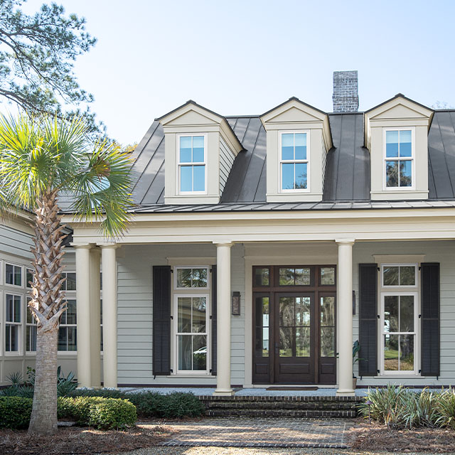 A beautiful gray-painted home features a white columned front porch and white trim, black shutters, a black glass-paned front door, dormer windows, and a palm tree in the front yard.