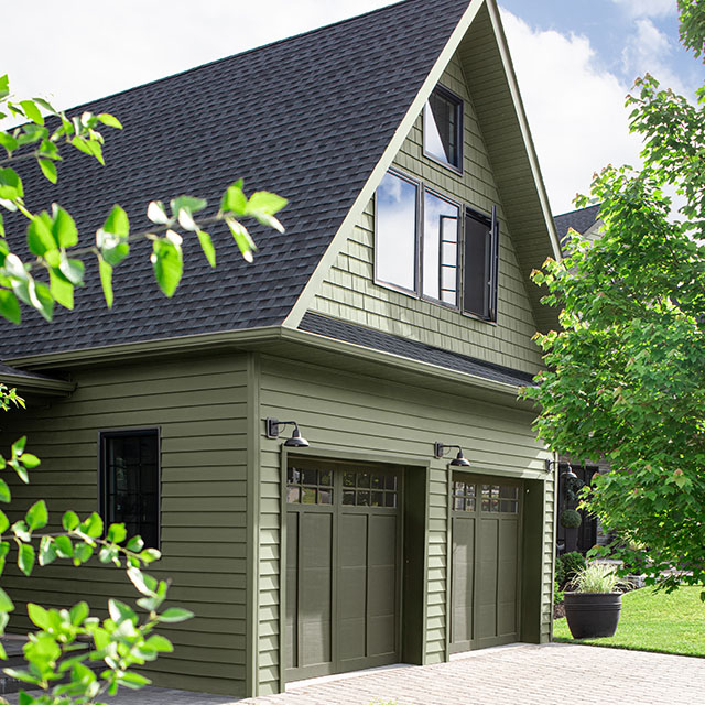 A partial home exterior with muted green-painted siding, second-story windows over mossy green-brown painted garage doors, and a green lawn with shrubs.