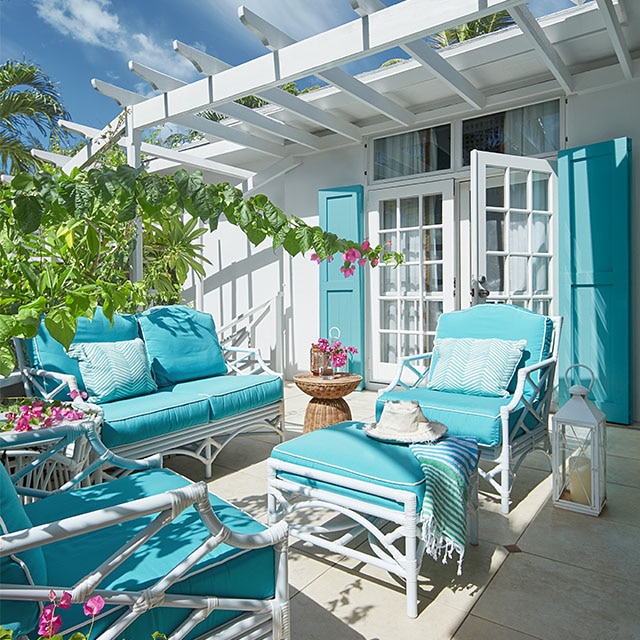 A pretty white-painted home exterior with turquoise shutters, an attached pergola and open french doors to a patio with turquoise cushioned furniture and tropical greenery.