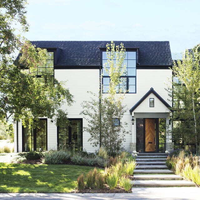 A beautiful home with white-painted siding on the left, stone exterior on the right side, and large black framed windows throughout, surrounded by greenery.