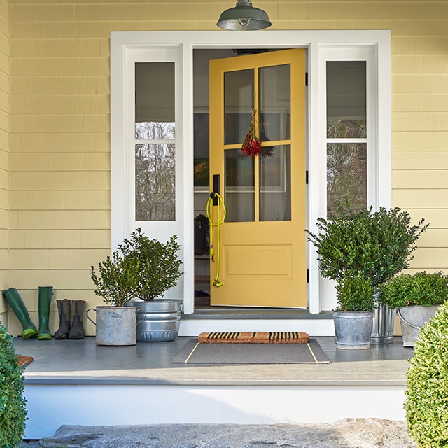 El porche frontal de una casa pintada de amarillo con una puerta amarilla, marco blanco, plantas en baldes de metal y dos arbustos redondos delante.