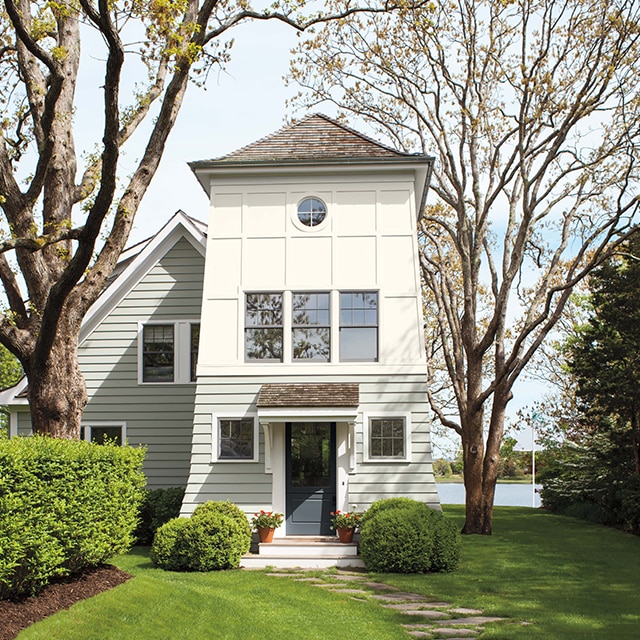 A charming lakeside two-tone painted house with off-white siding, white upper siding, a dark-gray front door, and lush lawn and greenery.