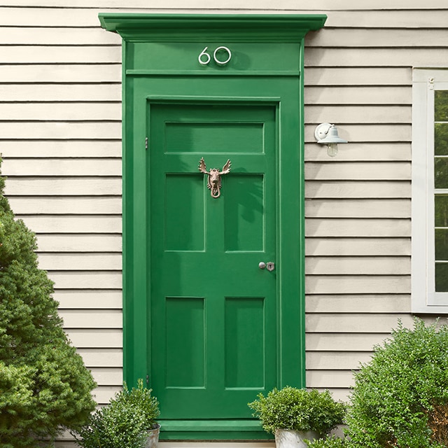 Warm greige-painted house siding, with a pretty green door flanked by shrubs, and white window trim.