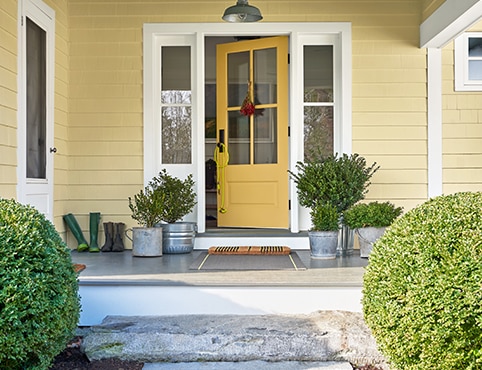 The front porch of a yellow-painted house with a yellow door, white trim, plants in metal pails, and two round shrubs out front.