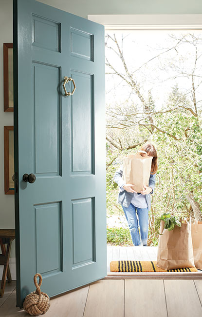 A soft blue-painted front door opens into a home as a woman carrying groceries walks towards it.
