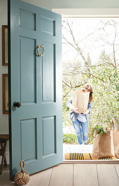 Una puerta principal pintada de azul suave se abre a una casa, mientras una mujer que lleva las compras camina hacia ella.