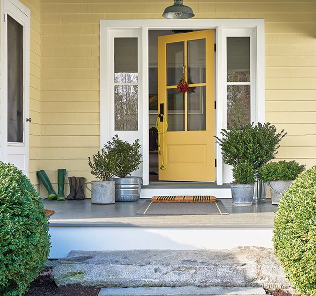 The front porch of a yellow-painted house with white trim, a yellow door, plants in metal pails, and two round shrubs out front.