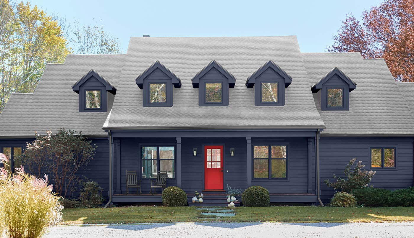 A beautiful Cape Cod house exterior painted in navy blue with a striking red door, surrounded by shrubberies, a green lawn, and a gravel driveway. 