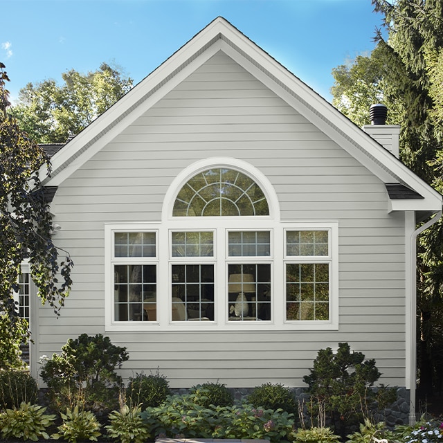 A house with light gray-painted exterior vinyl siding and white trimmed windows, surrounded by shrubs and trees.