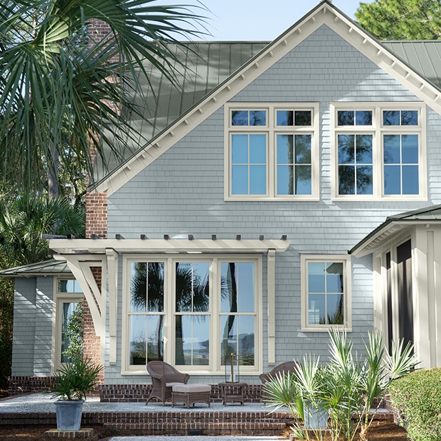 A stylish home with pale-blue painted siding with gray undertones, a silver-gray roof, and white window trim, surrounded by trees and a brick and concrete patio. 