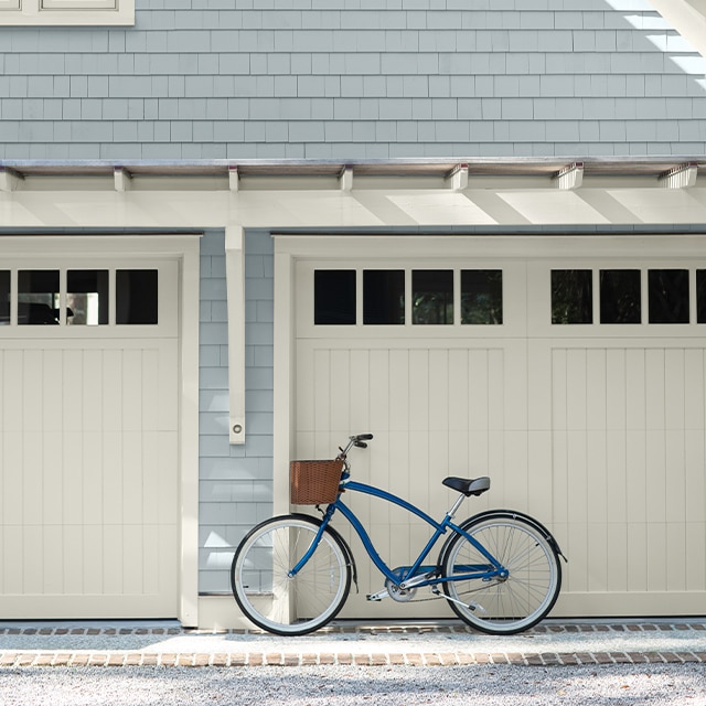 Pretty pale gray-painted home siding with a hint of blue-green, off-white trim, eaves and a blue bike leaning against one of two off-white garage doors.