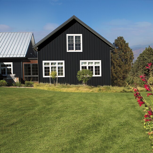 Maison noire de style ranch avec moulures de fenêtres blanches, pelouse et autres verdures luxuriantes.