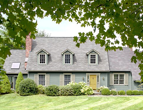 A beautiful cape style home with soft blue-gray painted siding, pale green shutters and trim, a yellow door, dormer windows, a lush green lawn and shrubbery.