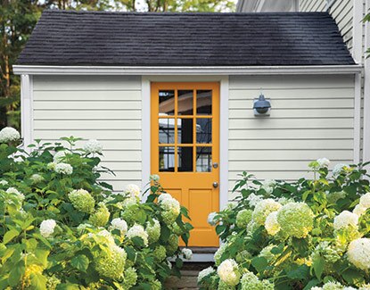 An off-white house with an orange-painted door and black roof with flowering green shrubbery.