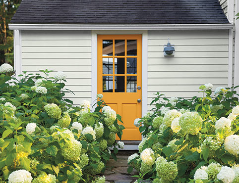 Cette maison blanc cassé avec porte d'entrée orange et toit noir est ornée d'arbustes fleuris.