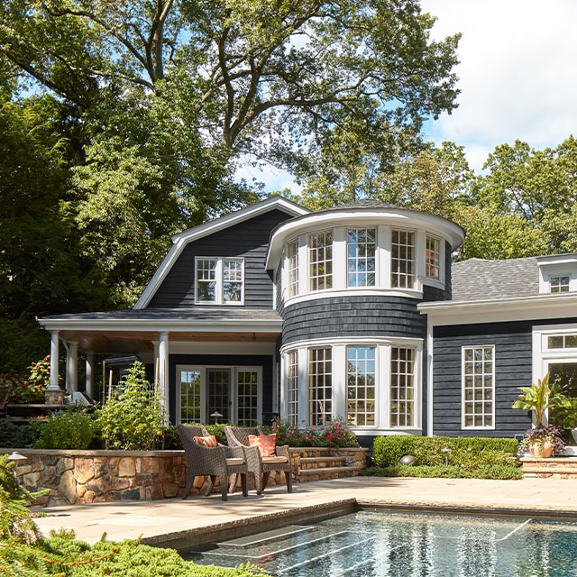 Una hermosa casa con revestimientos exteriores pintados de gris intenso y los marcos blancos, un porche esquinero con columnas blancas y un patio trasero con piscina.