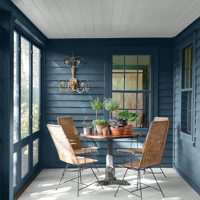Un porche cerrado, pintado de azul, con techo de listones blancos, piso blanco y una mesa redonda con plantas en macetas y sillas de mimbre.