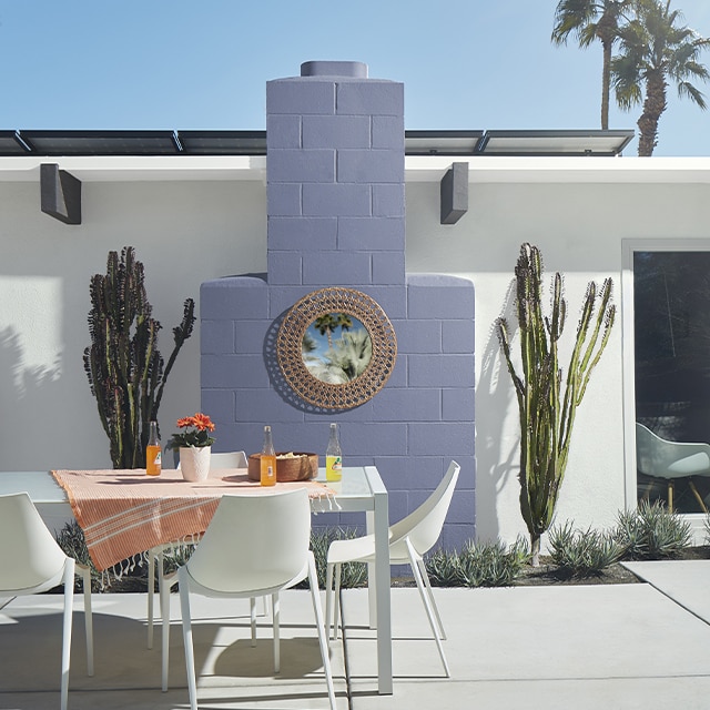Una casa de estuco pintada de blanco con una chimenea de color púrpura claro flanqueada por dos cactus convierte este patio con muebles de comedor blancos en un divertido espacio al aire libre.
