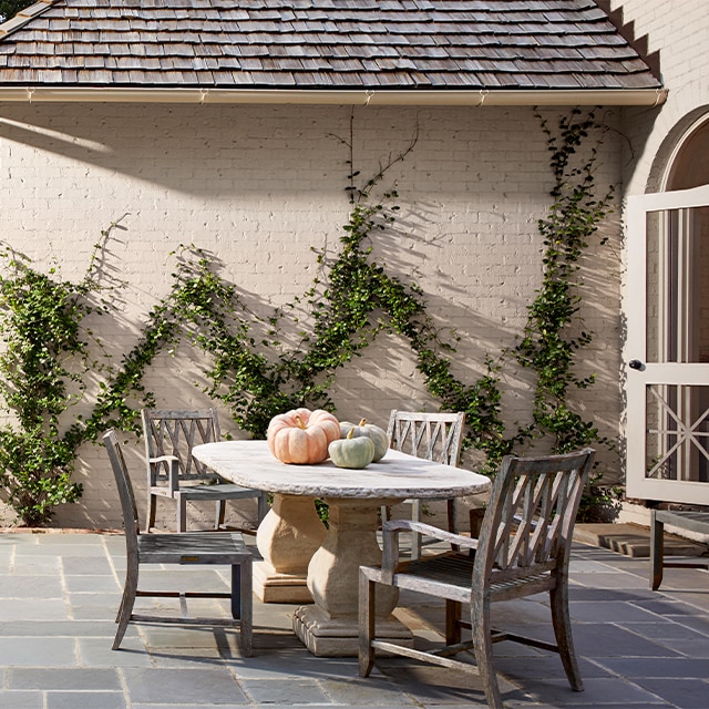 El exterior de una casa de ladrillos pintados de blanco con un suave matiz durazno y enredaderas trepadoras, alberga un espacioso comedor en el patio de pizarra con una mesa de piedra y sillas de madera.