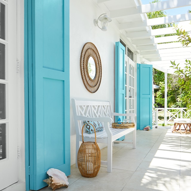 Un porche de ambiente tropical pintado de blanco con pérgola adosada, los postigos altos de color turquesa, un banco blanco y artículos decorativos de mimbre.