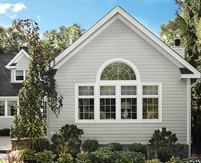 A house with light gray-painted exterior siding and white trimmed windows, surrounded by shrubs and trees.