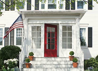 A white vinyl-sided home with a red window-paned front door and enclosed front portico welcomes guests. A white vinyl-sided home with a red window-paned front door and enclosed front portico welcomes guests.