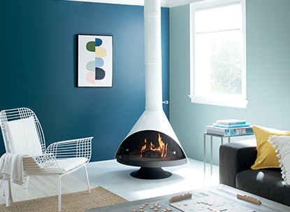 A living room with one light blue wall and one dark blue wall, modern white fireplace, wire frame chair, colourful wall art, and black sofa.