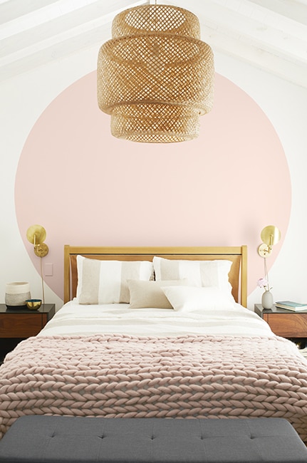 Light bedroom with white walls and a large light pink circle behind the bed with pink throw blanket and a basket chandelier.
