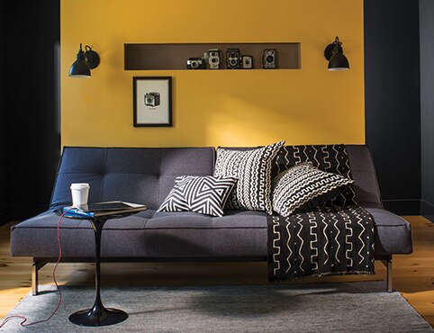 A contemporary living room with gray couch, graphic pillows and blankets, small end table, and yellow-painted accent wall.