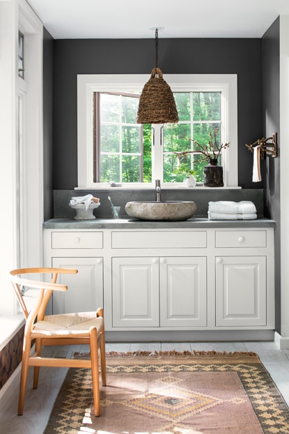 A rustic, yet modern, bathroom with stone sink on white cabinet, wooden chair, natural pendant lamp, and dark gray accent walls.