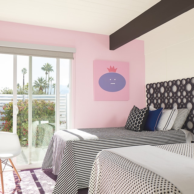 A bedroom with white-painted walls and ceiling, a cheery pink-painted accent wall, two twin beds with black and white polka dot and striped bedding, and a sliding door to a deck with views of palm trees and open sky.