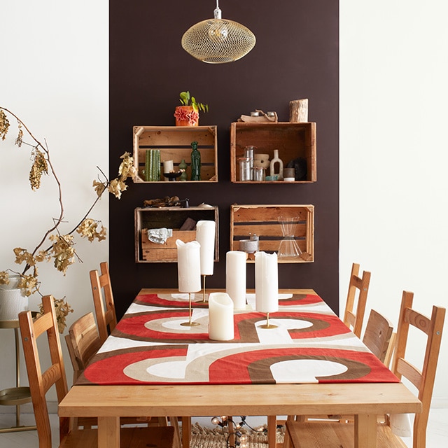 Comedor de apartamento moderno con un panel de acento pintado de marrón oscuro sobre una pared blanca, estanterías de pared con cajones, mesa y sillas de madera, y camino de mesa Naranja y marrón.