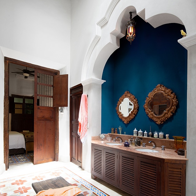 A pretty, white-painted bathroom with decorative moulding, a double wooden vanity set in a blue-painted alcove, a colourful tile floor, wood bench and doors opening to a bedroom.