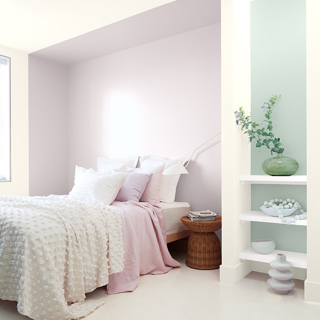 A white bedroom with a violet painted inset behind a bed with white and pink bedding, and a blue-green accent wall behind white shelving.