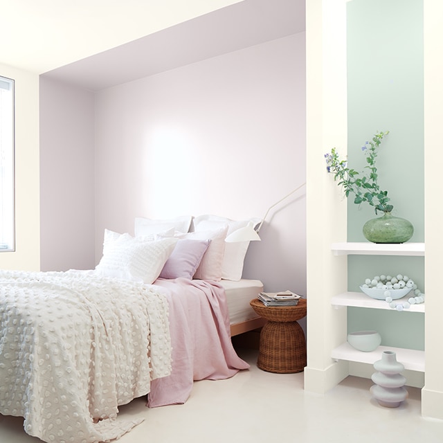 Una habitación blanca con un estante pintado de violeta detrás de una cama con ropa de cama blanca y rosa, y una pared de acento azul verdoso detrás de estanterías blancas.