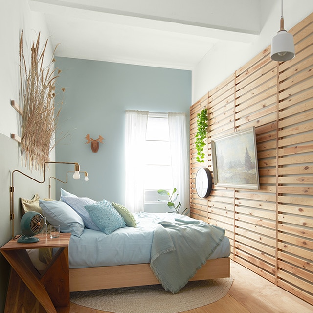A cozy, sunlit bedroom with a light blue-painted accent wall, a two-tone muted silver and white wall behind a bed, a wood slatted wall with hanging artwork and a raised wood floor.