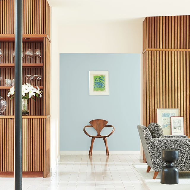 Una sala de estar luminosa y abierta con paneles y gabinetes acanalados de madera, paredes blancas, una silla negra y una moderna silla de madera contra una pared del pasillo pintada de azul claro.