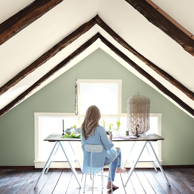 A woman sits at a contemporary desk in a small office space with a sloped white ceiling with wooden beams and a soft green-painted accent wall framing windows.