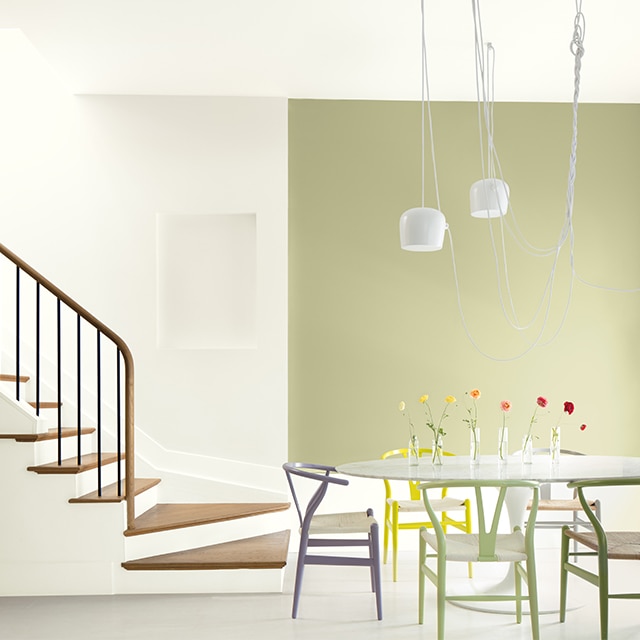 An open dining area with a light-green painted accent wall, white walls and ceiling, multi-coloured chairs and a white table near a white open staircase with wood steps and banister.