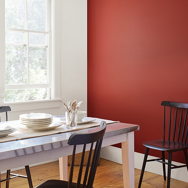 A dining room with a red-painted accent wall, a white wall, trim and ceiling, wood floors, a white table set with white dishes, and black spindle-back chairs.