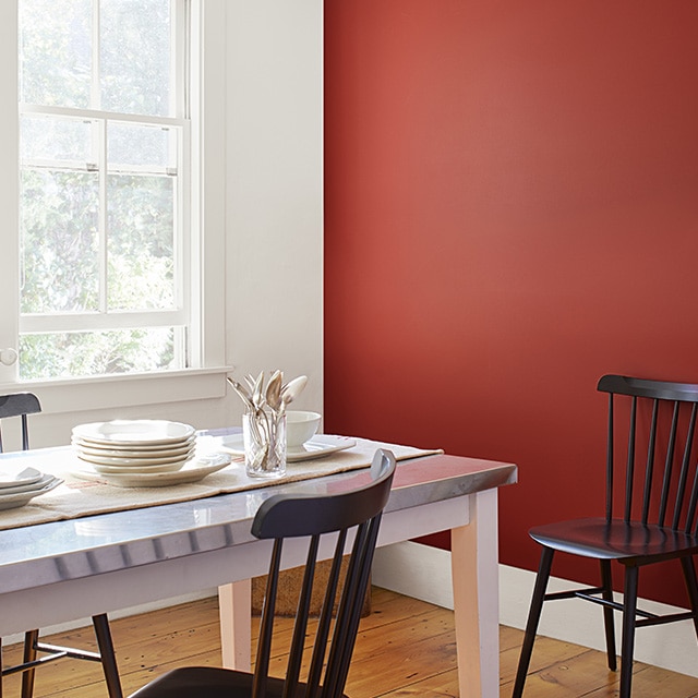 Un comedor con una pared de acento pintada de rojo, otra pared, marcos y techo en blanco, piso de madera, una mesa blanca con vajilla blanca y sillas negras con respaldo de barrotes.