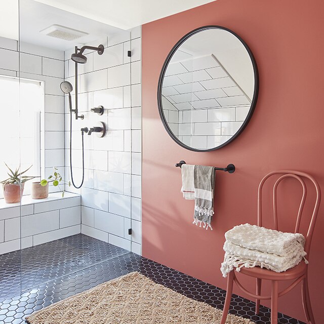 A bathroom with a red-painted accent wall, round mirror with black frame, a red-painted chair holding towels, white tiled walls and black tiled flooring with a beige bathmat.