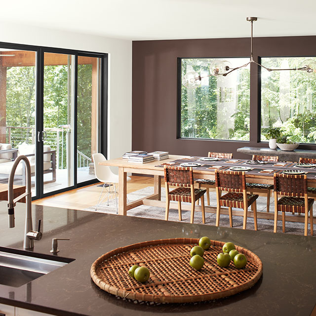 An open, modern dining room with a brown-painted accent wall and three large windows, a white ceiling and side wall with sliding glass doors, and brown granite kitchen island in the forefront.