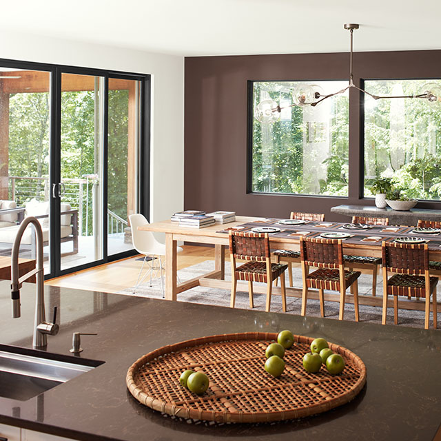 An open, modern dining room with a brown-painted accent wall and three large windows, a white ceiling and side wall with sliding glass doors, and brown granite kitchen island in the forefront. 