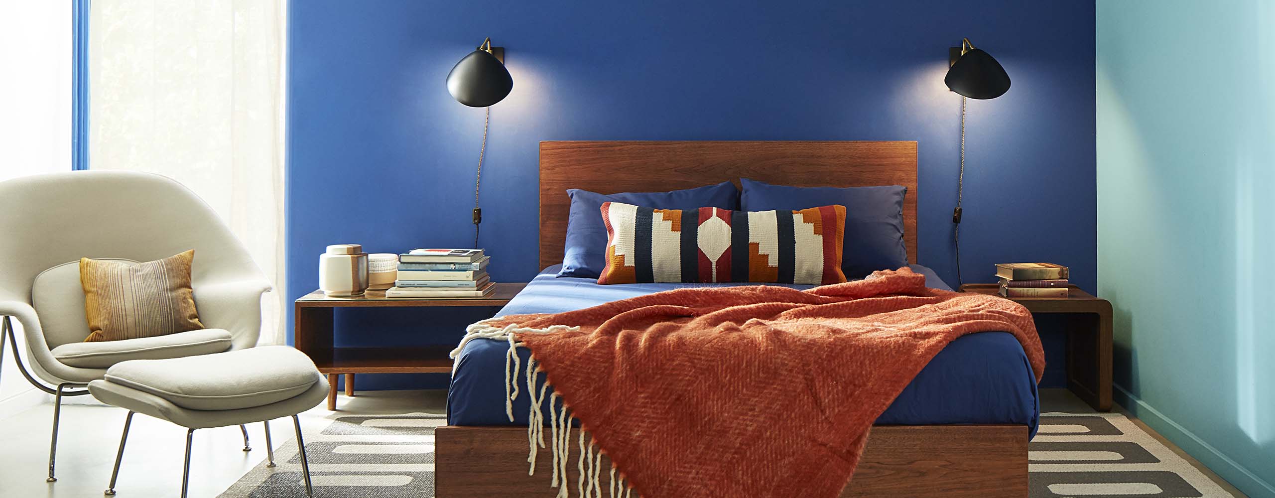 A bedroom with two blue-painted accent walls, a wooden bed frame, rust coloured throw, contemporary beige chair, and reading lamps on both sides of bed.