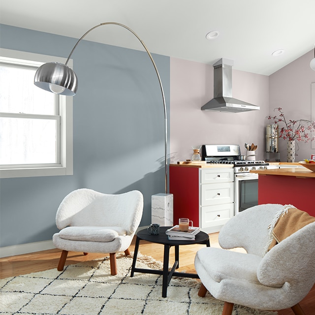 A bright, open apartment space with a light blue-painted living room wall, pale gray kitchen walls with a hint of lavender, a white vaulted ceiling, a deep red kitchen island, white kitchen drawers, and black and white modern decor. 