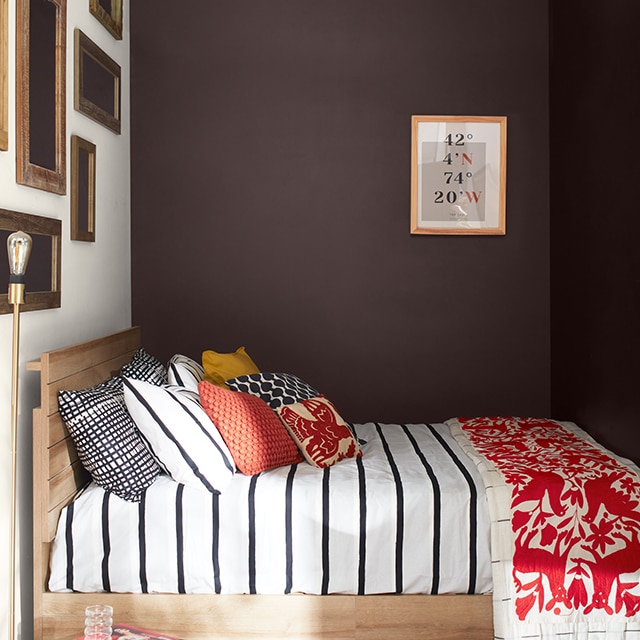 A cozy bedroom with deep chocolate-hued walls, a white-painted gallery wall, a bed with black and white striped bedding, pillows and red printed blanket, and a wicker ottoman.